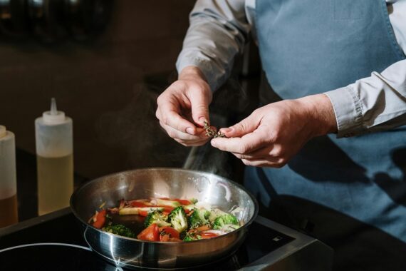 Chef hands adding fresh thyme herbs to a hot skillet of sautéed vegetables, demonstrating late-stage spice timing for maximum flavor and freshness in cooking.